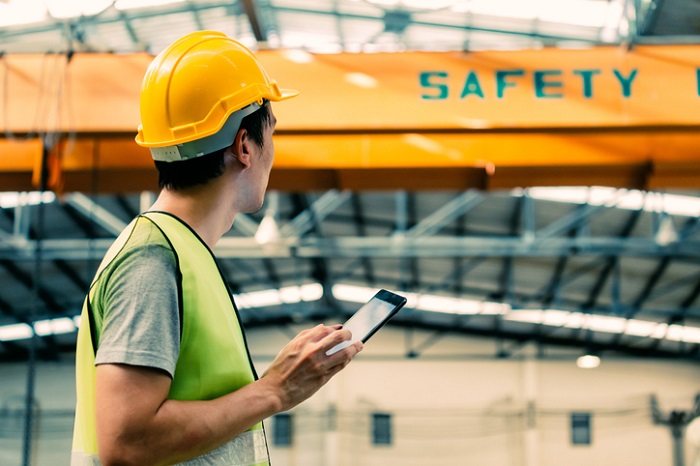 Mann mit Arbeitshelm auf dem Kopf und Tablet in der Hand steht in einer Arbeitshalle